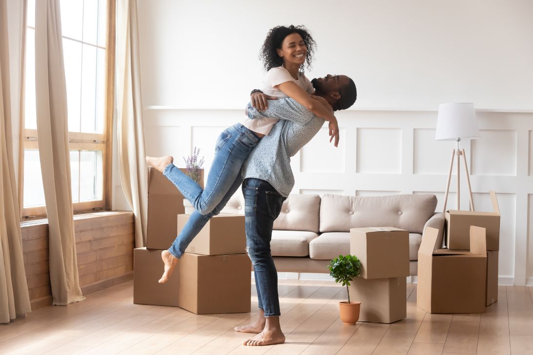 A man is lifting a woman off the ground in a celebratory manner with moving boxes in the background as if they are first time homebuyers during a recession but succeeded in buying a home