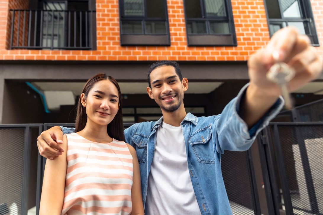 A man and woman are smiling and holding out a key as the decided to buy a home after learning is buying a home during a recession worth it