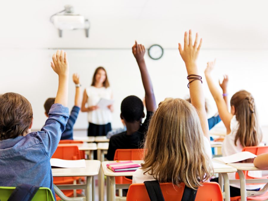 Students in a classroom with their hands raised looking at teacher in the foreground illustrative of what you would see in Cedar Lake Indiana Schools